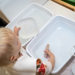 Toddler washing dishes in white plastic tubs.